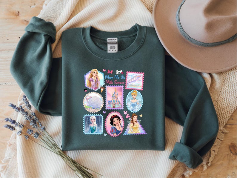 May include: Dark green sweatshirt featuring a colorful graphic of various Disney princesses designed as postage stamps. The text "Meet Me On Main Street" is also included. The sweatshirt has long sleeves and a crew neck.