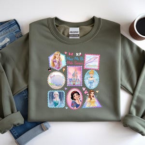 May include: An olive green sweatshirt featuring a colorful graphic of various Disney princesses and landmarks, designed in a postage stamp style. The graphic includes the text "Meet Me at the Sweet Spot". The sweatshirt is paired with blue jeans.