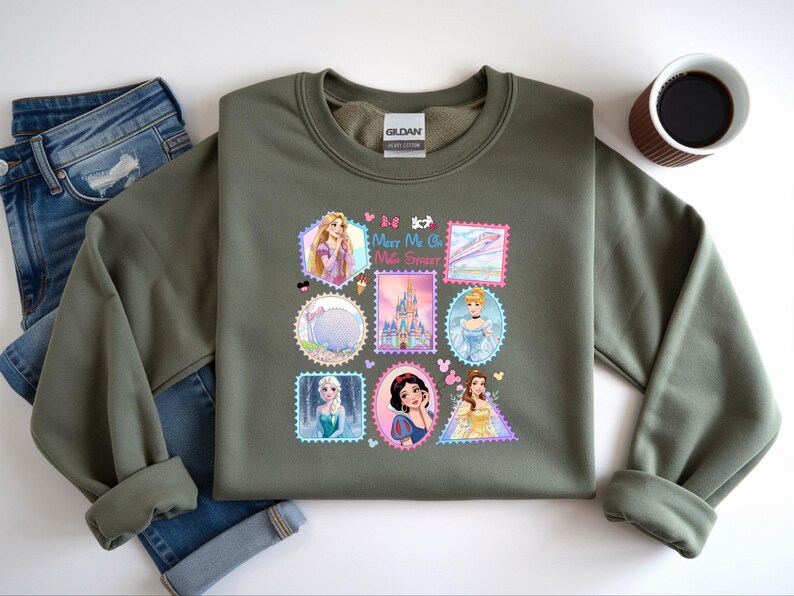 May include: An olive green sweatshirt featuring a colorful graphic of various Disney princesses and landmarks, designed in a postage stamp style. The graphic includes the text "Meet Me at the Sweet Spot". The sweatshirt is paired with blue jeans.