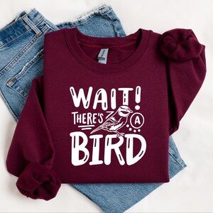 May include: A maroon sweatshirt with the text "WAIT! THERE'S A BIRD" in white lettering. The graphic includes a stylized bird with a white outline.