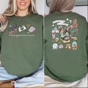 以下が含まれることがあります： ピンクのリボンが付いたミッキーマウスの耳や「The Most Magical Place On Earth」の文字など、ディズニーをテーマにしたグラフィックが特徴のオリーブグリーンのスウェットシャツ。背面には、さらに多くのディズニーのキャラクターやランドマークが描かれています。