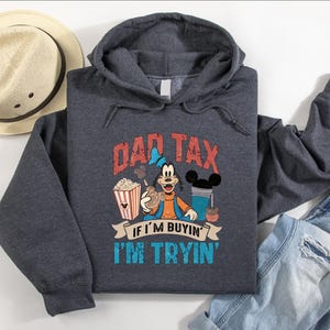 Pode incluir: Uma camisola com capuz cinzento-escuro mesclado com um gr&aacute;fico da personagem Pateta segurando um balde de pipocas e uma bebida. O texto diz DAD TAX IF I'M BUYIN' I'M TRYIN'. A camisola &eacute; exibida ao lado de um chap&eacute;u de palha e cal&ccedil;as de ganga azuis.