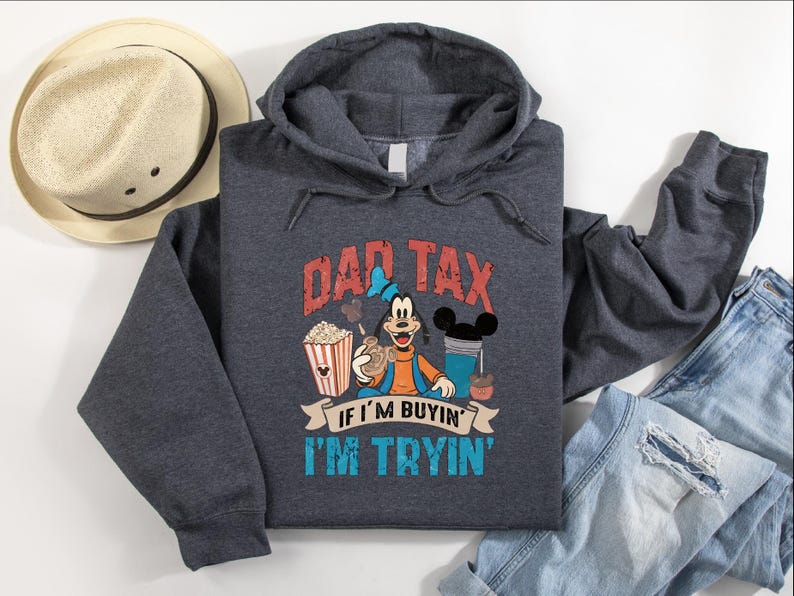 Pode incluir: Uma camisola com capuz cinzento-escuro mesclado com um gr&aacute;fico da personagem Pateta segurando um balde de pipocas e uma bebida. O texto diz DAD TAX IF I'M BUYIN' I'M TRYIN'. A camisola &eacute; exibida ao lado de um chap&eacute;u de palha e cal&ccedil;as de ganga azuis.