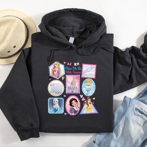 May include: Black hooded sweatshirt featuring a colorful graphic of postage stamp designs with Disney princesses. The stamps include images of Rapunzel, Cinderella, Snow White, and Belle. The text "Meet Me On Main Street" is also included.