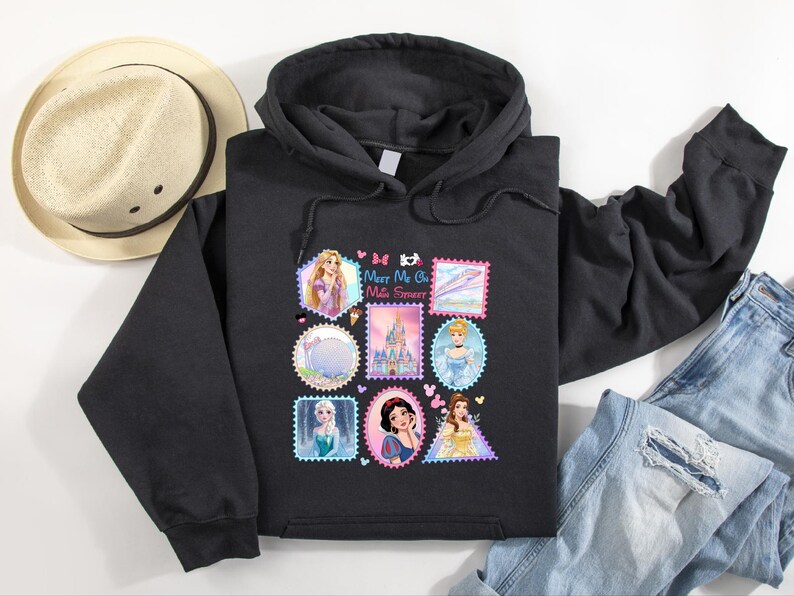 May include: Black hooded sweatshirt featuring a colorful graphic of postage stamp designs with Disney princesses. The stamps include images of Rapunzel, Cinderella, Snow White, and Belle. The text "Meet Me On Main Street" is also included.