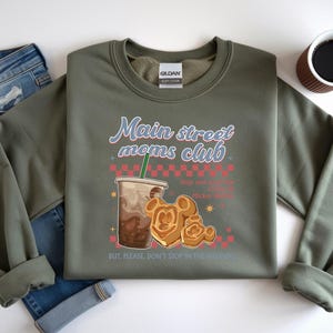 Op de afbeelding: Olijfgroene sweatshirt met de tekst "Main Street Moms Club" in een retro lettertype. Het ontwerp bevat een koffiedrankje, Mickey Mouse-wafels en de zin "Stop and smell the Mickey Waffles". Een spijkerbroek en een kopje koffie staan ook op de afbeelding.