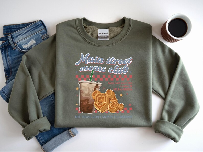 Op de afbeelding: Olijfgroene sweatshirt met de tekst "Main Street Moms Club" in een retro lettertype. Het ontwerp bevat een koffiedrankje, Mickey Mouse-wafels en de zin "Stop and smell the Mickey Waffles". Een spijkerbroek en een kopje koffie staan ook op de afbeelding.