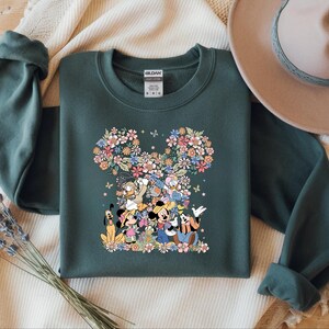 May include: Dark green sweatshirt featuring a floral Mickey Mouse head design. The design incorporates various Disney characters and colorful flowers. A tan hat and a sprig of lavender are in the background.