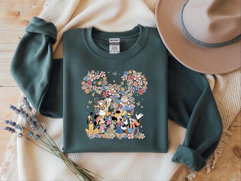 May include: Dark green sweatshirt featuring a floral Mickey Mouse head design. The design incorporates various Disney characters and colorful flowers. A tan hat and a sprig of lavender are in the background.