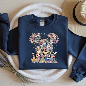 May include: Navy blue sweatshirt featuring a floral Mickey Mouse design. The design includes Mickey, Minnie, Donald, Goofy, and other characters. The floral design is in shades of pink, yellow, and blue. The sweatshirt has long sleeves and a crew neck.