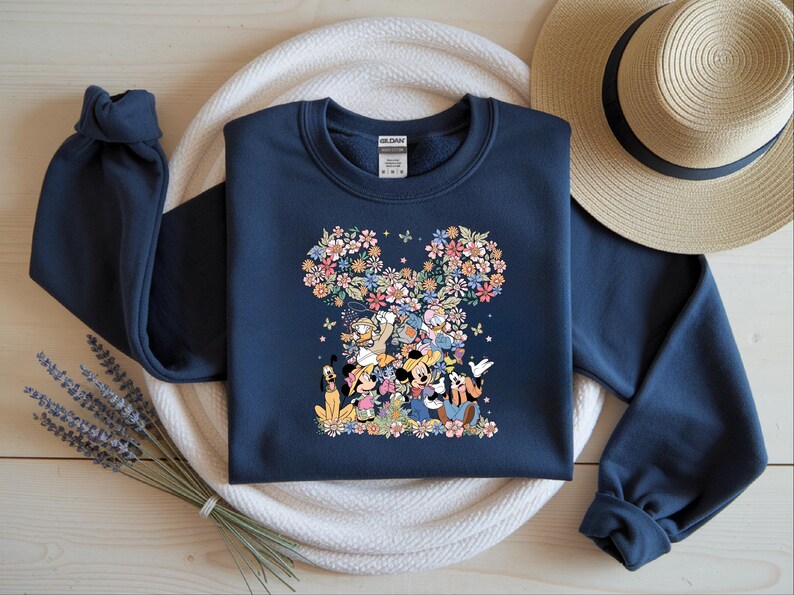 May include: Navy blue sweatshirt featuring a floral Mickey Mouse design. The design includes Mickey, Minnie, Donald, Goofy, and other characters. The floral design is in shades of pink, yellow, and blue. The sweatshirt has long sleeves and a crew neck.