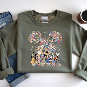 May include: An olive green sweatshirt featuring a floral Mickey Mouse design with various Disney characters. The design includes Mickey, Minnie, Donald, Goofy, and Pluto, surrounded by colorful flowers. The sweatshirt is paired with blue jeans and a cup of coffee.