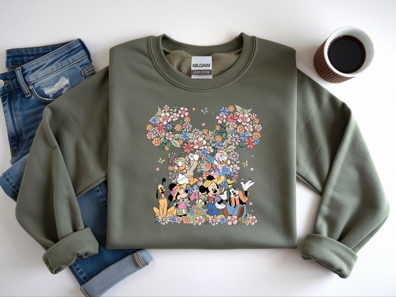 May include: An olive green sweatshirt featuring a floral Mickey Mouse design with various Disney characters. The design includes Mickey, Minnie, Donald, Goofy, and Pluto, surrounded by colorful flowers. The sweatshirt is paired with blue jeans and a cup of coffee.