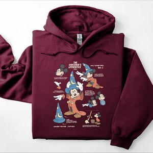 Puede incluir: Una sudadera con capucha de color granate que presenta un estampado gr&aacute;fico de El Aprendiz de Brujo de Fantas&iacute;a. El dise&ntilde;o incluye varias ilustraciones de Mickey Mouse con sombrero de mago, junto con notas t&eacute;cnicas y texto descriptivo.