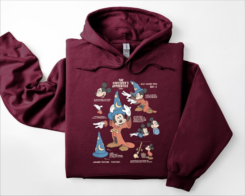 Puede incluir: Una sudadera con capucha de color granate que presenta un estampado gr&aacute;fico de El Aprendiz de Brujo de Fantas&iacute;a. El dise&ntilde;o incluye varias ilustraciones de Mickey Mouse con sombrero de mago, junto con notas t&eacute;cnicas y texto descriptivo.