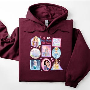May include: A maroon hooded sweatshirt featuring a colorful graphic of Disney princesses and landmarks in a postage stamp design. The text "Meet Me On Main Street" is also visible on the front of the sweatshirt.