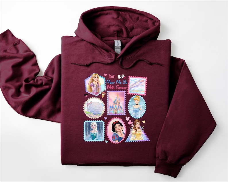 May include: A maroon hooded sweatshirt featuring a colorful graphic of Disney princesses and landmarks in a postage stamp design. The text "Meet Me On Main Street" is also visible on the front of the sweatshirt.