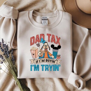 Pode incluir: Uma camisola de gola redonda bege com um gr&aacute;fico da personagem de desenhos animados Pateta rodeada de pipocas, uma bebida e snacks. O texto DAD TAX IF I'M BUYIN' I'M TRYIN' est&aacute; escrito em letras vermelhas e azuis. Etiqueta Gildan vis&iacute;vel.
