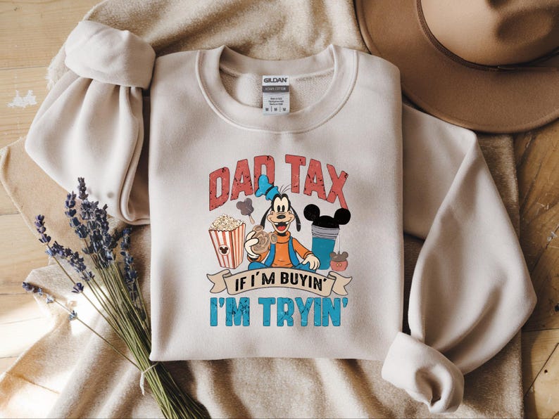 Pode incluir: Uma camisola de gola redonda bege com um gr&aacute;fico da personagem de desenhos animados Pateta rodeada de pipocas, uma bebida e snacks. O texto DAD TAX IF I'M BUYIN' I'M TRYIN' est&aacute; escrito em letras vermelhas e azuis. Etiqueta Gildan vis&iacute;vel.