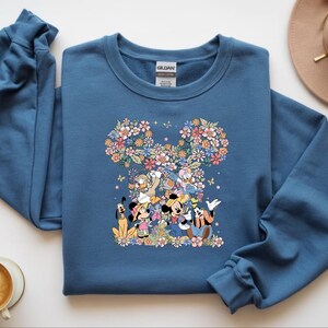 May include: A blue sweatshirt featuring a floral Mickey Mouse design with various Disney characters. The design includes Mickey, Minnie, Donald, Goofy, and Pluto, surrounded by colorful flowers and butterflies. A tan hat and a cup of coffee are also visible.