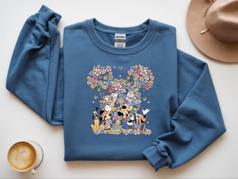 May include: A blue sweatshirt featuring a floral Mickey Mouse design with various Disney characters. The design includes Mickey, Minnie, Donald, Goofy, and Pluto, surrounded by colorful flowers and butterflies. A tan hat and a cup of coffee are also visible.