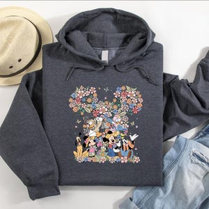 May include: A dark gray hooded sweatshirt featuring a floral Mickey Mouse design. The design includes various Disney characters and colorful flowers. A straw hat and blue jeans are also visible.