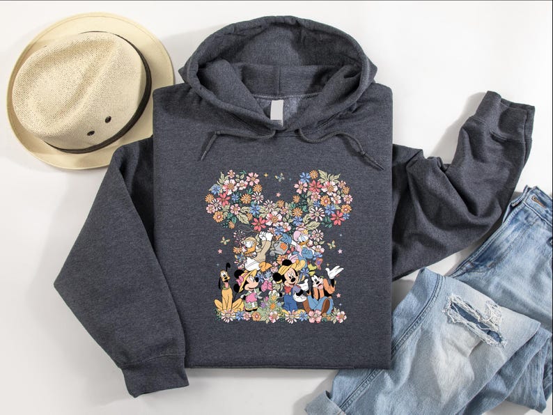 May include: A dark gray hooded sweatshirt featuring a floral Mickey Mouse design. The design includes various Disney characters and colorful flowers. A straw hat and blue jeans are also visible.