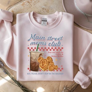 Op de afbeelding: Een lichtroze sweatshirt met de tekst "Main street moms club" en een afbeelding van koffie en Mickey Mouse wafels. De sweatshirt heeft lange mouwen en een ronde hals.