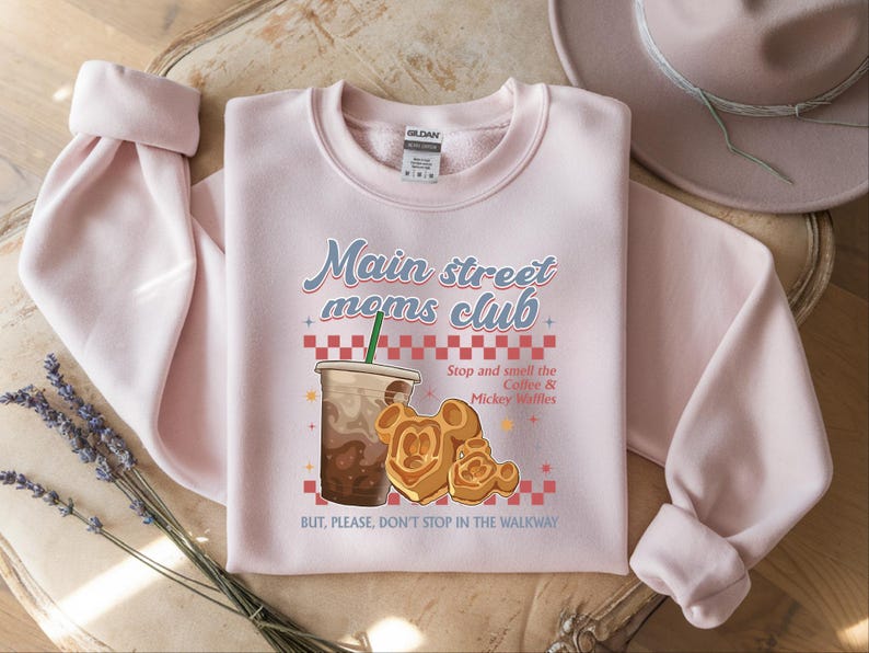 Op de afbeelding: Een lichtroze sweatshirt met de tekst "Main street moms club" en een afbeelding van koffie en Mickey Mouse wafels. De sweatshirt heeft lange mouwen en een ronde hals.