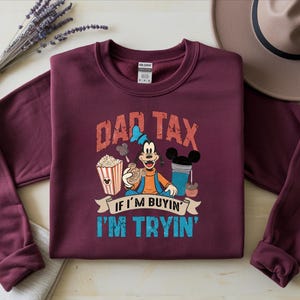 Pode incluir: Uma camisola de manga comprida cor de vinho com um gr&aacute;fico da personagem Pateta a segurar um snack, rodeado de pipocas e uma bebida. O texto DAD TAX IF I'M BUYIN' I'M TRYIN' est&aacute; em letras vermelhas e azuis gastas na parte da frente.