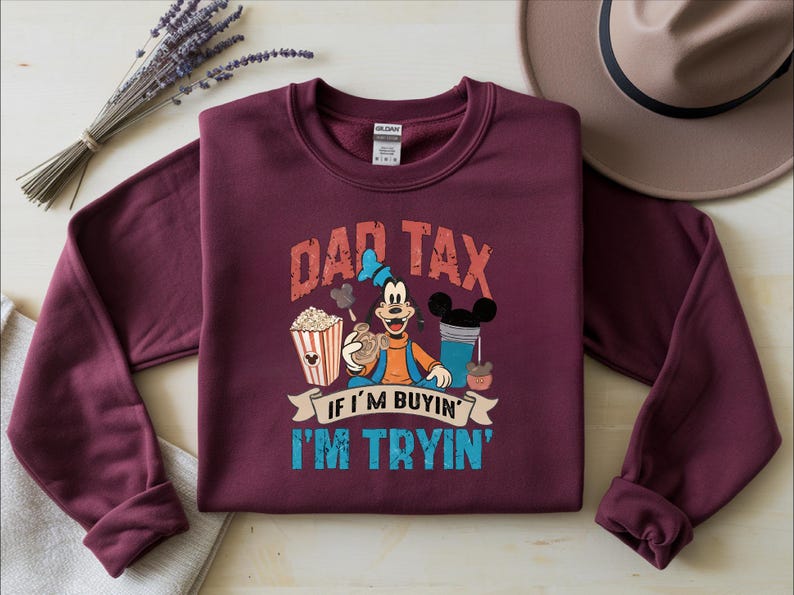 Pode incluir: Uma camisola de manga comprida cor de vinho com um gr&aacute;fico da personagem Pateta a segurar um snack, rodeado de pipocas e uma bebida. O texto DAD TAX IF I'M BUYIN' I'M TRYIN' est&aacute; em letras vermelhas e azuis gastas na parte da frente.