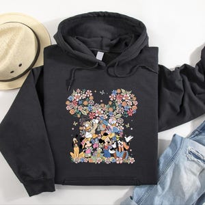 May include: Black hooded sweatshirt featuring a floral Mickey Mouse design. The design incorporates various characters and colorful flowers. A straw hat and blue jeans are also visible.