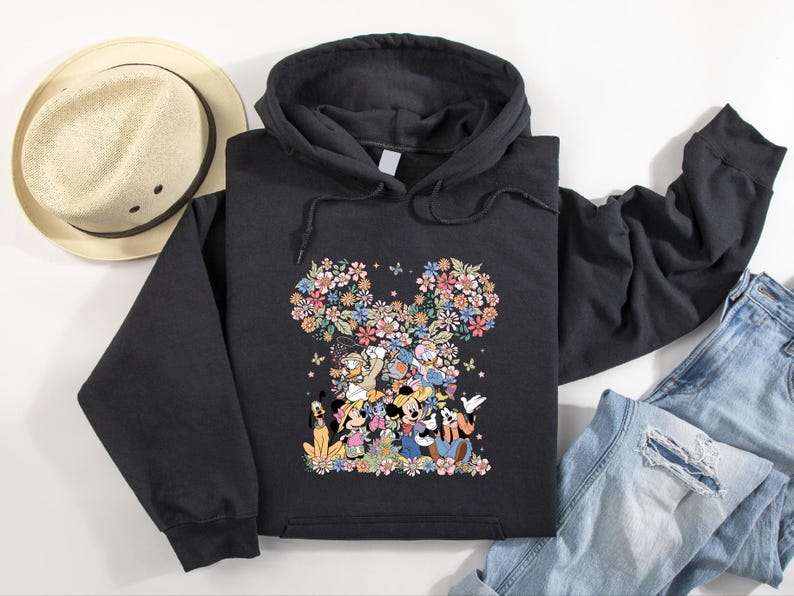 May include: Black hooded sweatshirt featuring a floral Mickey Mouse design. The design incorporates various characters and colorful flowers. A straw hat and blue jeans are also visible.
