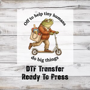 May include: DTF transfer featuring a cartoon frog riding a scooter, wearing a sweater and carrying a tote bag. The text reads "Off to help tiny humans do big things" and "DTF Transfer Ready To Press". The frog is green, the scooter is silver.