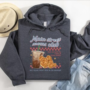Op de afbeelding: Een donkergrijze hoodie met de tekst "Main Street Moms Club" en een illustratie van koffie en Mickey Mouse wafels. De hoodie heeft ook de tekst "Stop and smell the Coffee & Mickey Waffles" en "BUT, PLEASE, DON'T STOP IN THE WALKWAY". Een strohoed en jeans staan ook op de foto.