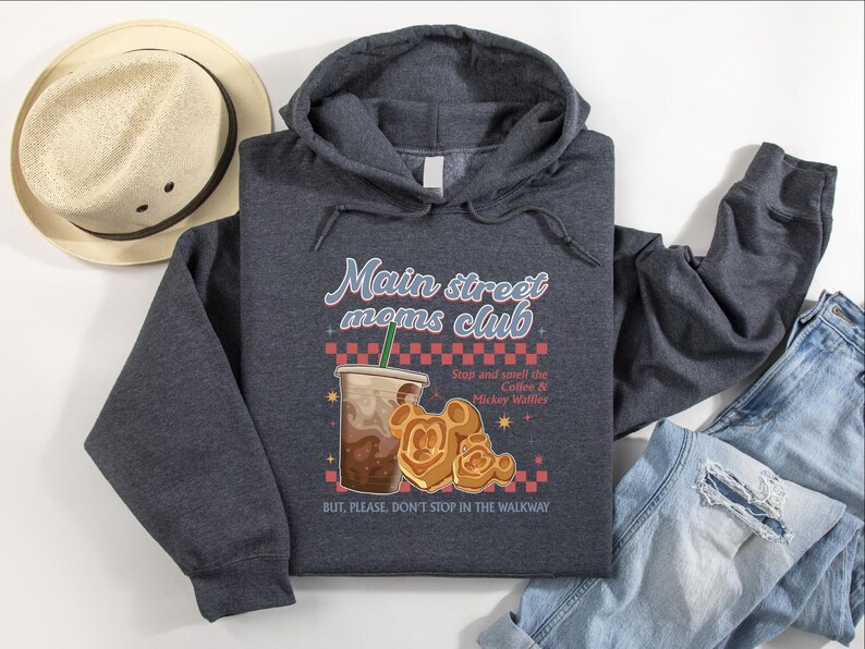 Op de afbeelding: Een donkergrijze hoodie met de tekst "Main Street Moms Club" en een illustratie van koffie en Mickey Mouse wafels. De hoodie heeft ook de tekst "Stop and smell the Coffee & Mickey Waffles" en "BUT, PLEASE, DON'T STOP IN THE WALKWAY". Een strohoed en jeans staan ook op de foto.