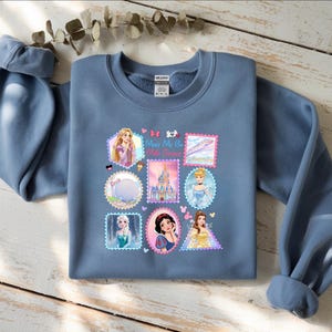 May include: A blue sweatshirt featuring a collage of Disney princess images in postage stamp designs. The sweatshirt has long sleeves and a crew neck. The text "Merry Me" is visible on the design. The sweatshirt is laying on a wooden surface.