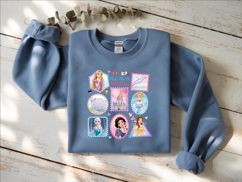 May include: A blue sweatshirt featuring a collage of Disney princess images in postage stamp designs. The sweatshirt has long sleeves and a crew neck. The text "Merry Me" is visible on the design. The sweatshirt is laying on a wooden surface.