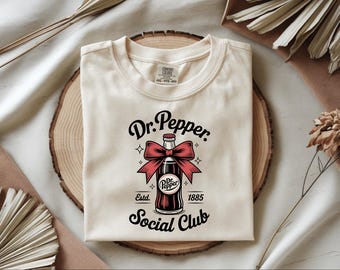 May include: Cream-colored t-shirt featuring a Dr. Pepper bottle design with a red bow, stars, and the words "Dr. Pepper Social Club" and "Estd. 1885." The shirt is folded on a wooden surface.