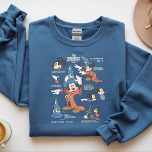 Puede incluir: Una sudadera azul de cuello redondo con un estampado gr&aacute;fico de Mickey Mouse como el aprendiz de brujo. El dise&ntilde;o incluye varios bocetos del personaje, un sombrero de mago azul con una luna y estrellas, y texto con notas de animaci&oacute;n.