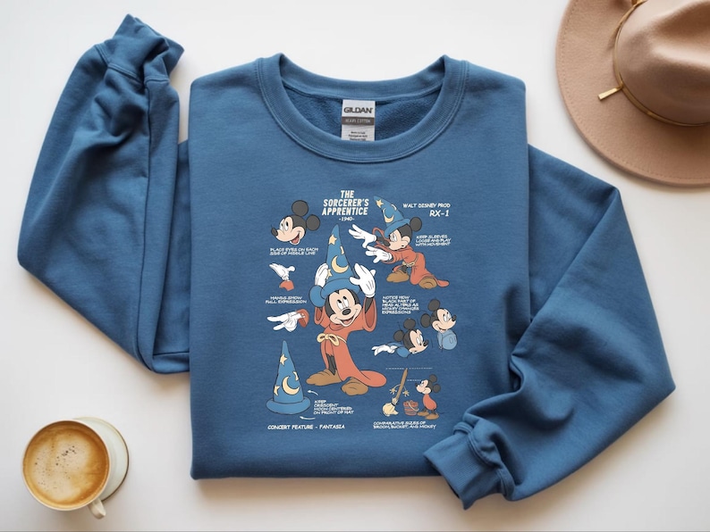 Puede incluir: Una sudadera azul de cuello redondo con un estampado gr&aacute;fico de Mickey Mouse como el aprendiz de brujo. El dise&ntilde;o incluye varios bocetos del personaje, un sombrero de mago azul con una luna y estrellas, y texto con notas de animaci&oacute;n.