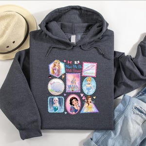 May include: A charcoal gray hooded sweatshirt featuring a colorful graphic of postage stamp-style images of Disney princesses. The sweatshirt includes the text "Meet Me at the Castle". A straw hat and blue jeans are also in the image.