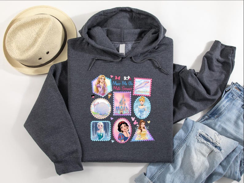 May include: A charcoal gray hooded sweatshirt featuring a colorful graphic of postage stamp-style images of Disney princesses. The sweatshirt includes the text "Meet Me at the Castle". A straw hat and blue jeans are also in the image.