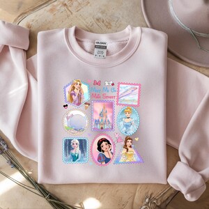 May include: A light pink sweatshirt featuring a collection of Disney princess postage stamp designs. The stamps include images of Rapunzel, Cinderella, Elsa, Snow White, and Belle. The text "Be My Oh Mile Street" is also visible on the sweatshirt.