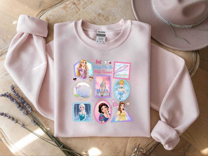 May include: A light pink sweatshirt featuring a collection of Disney princess postage stamp designs. The stamps include images of Rapunzel, Cinderella, Elsa, Snow White, and Belle. The text "Be My Oh Mile Street" is also visible on the sweatshirt.