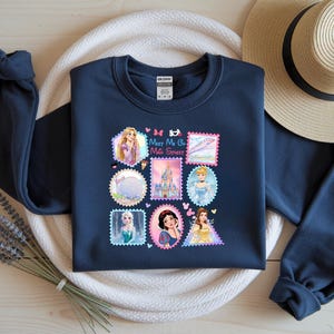 May include: Navy blue sweatshirt featuring a collection of princess-themed postage stamp designs. The stamps include images of princesses, castles, and other related graphics. The text "Meet Me at the Sweetest" is also included.