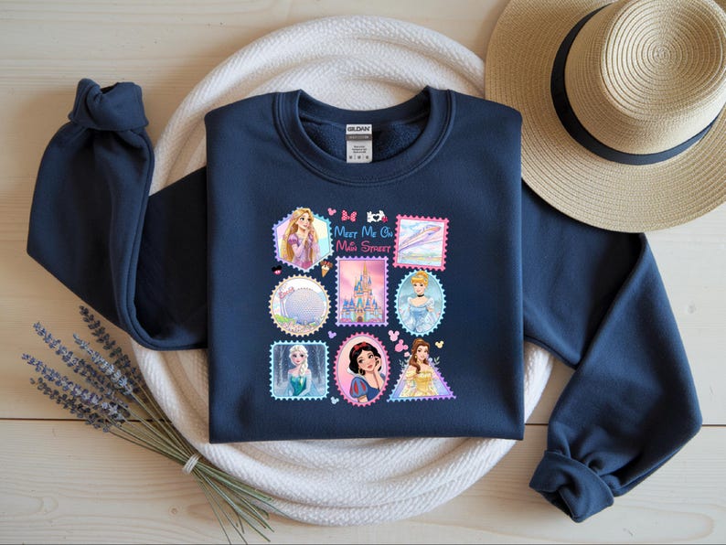 May include: Navy blue sweatshirt featuring a collection of princess-themed postage stamp designs. The stamps include images of princesses, castles, and other related graphics. The text "Meet Me at the Sweetest" is also included.