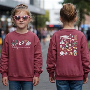 以下が含まれることがあります： ミッキーマウスの耳、ウサギ、そして「The Most Magical Place on Earth」の文字が描かれた、前面にグラフィックが施されたマルーン色のスウェットシャツ。スウェットシャツの背面には、さまざまなディズニーキャラクターのイラストが描かれています。子供はピンクのサングラスとブルージーンズを着用しています。