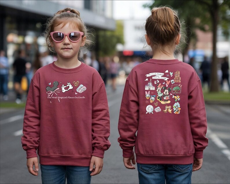 以下が含まれることがあります： ミッキーマウスの耳、ウサギ、そして「The Most Magical Place on Earth」の文字が描かれた、前面にグラフィックが施されたマルーン色のスウェットシャツ。スウェットシャツの背面には、さまざまなディズニーキャラクターのイラストが描かれています。子供はピンクのサングラスとブルージーンズを着用しています。
