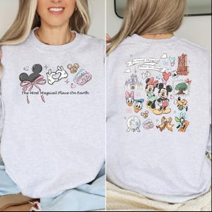 以下が含まれることがあります： ミッキーマウスの耳、ハート型の手、プレッツェルが描かれたライトグレーのスウェットシャツ。「The Most Magical Place On Earth」の文字が前面にプリントされています。背面には、ミッキーとミニーマウスを含む、さまざまなディズニーキャラクターのイラストが描かれています。